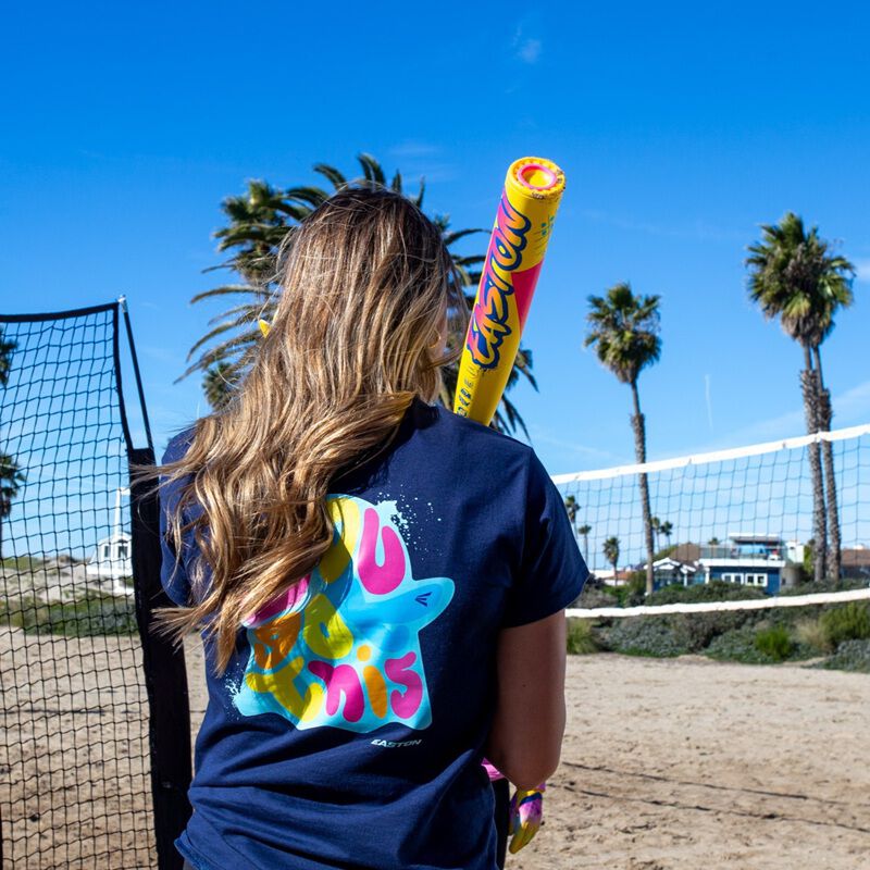 A person with long hair in a colorful t-shirt stands on a sandy beach holding the 2026 Easton Ghost Advanced Spring Break Yellow (-10) Fastpitch Softball Bat (EFP6GHSY10) by Easton, facing a beach volleyball net with palm trees and blue sky behind.