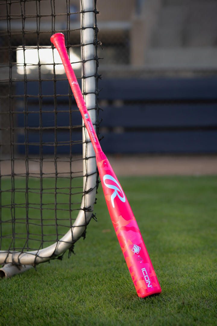 A 2025 Rawlings Icon Electric Unicorn (-3) BBCOR Baseball Bat (RBB5IPK3) in bright pink with white and red accents leans against a net on grass, blue benches and a fence visible in the background.
