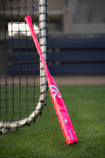 A 2025 Rawlings Icon Electric Unicorn (-3) BBCOR Baseball Bat (RBB5IPK3) in bright pink with white and red accents leans against a net on grass, blue benches and a fence visible in the background.