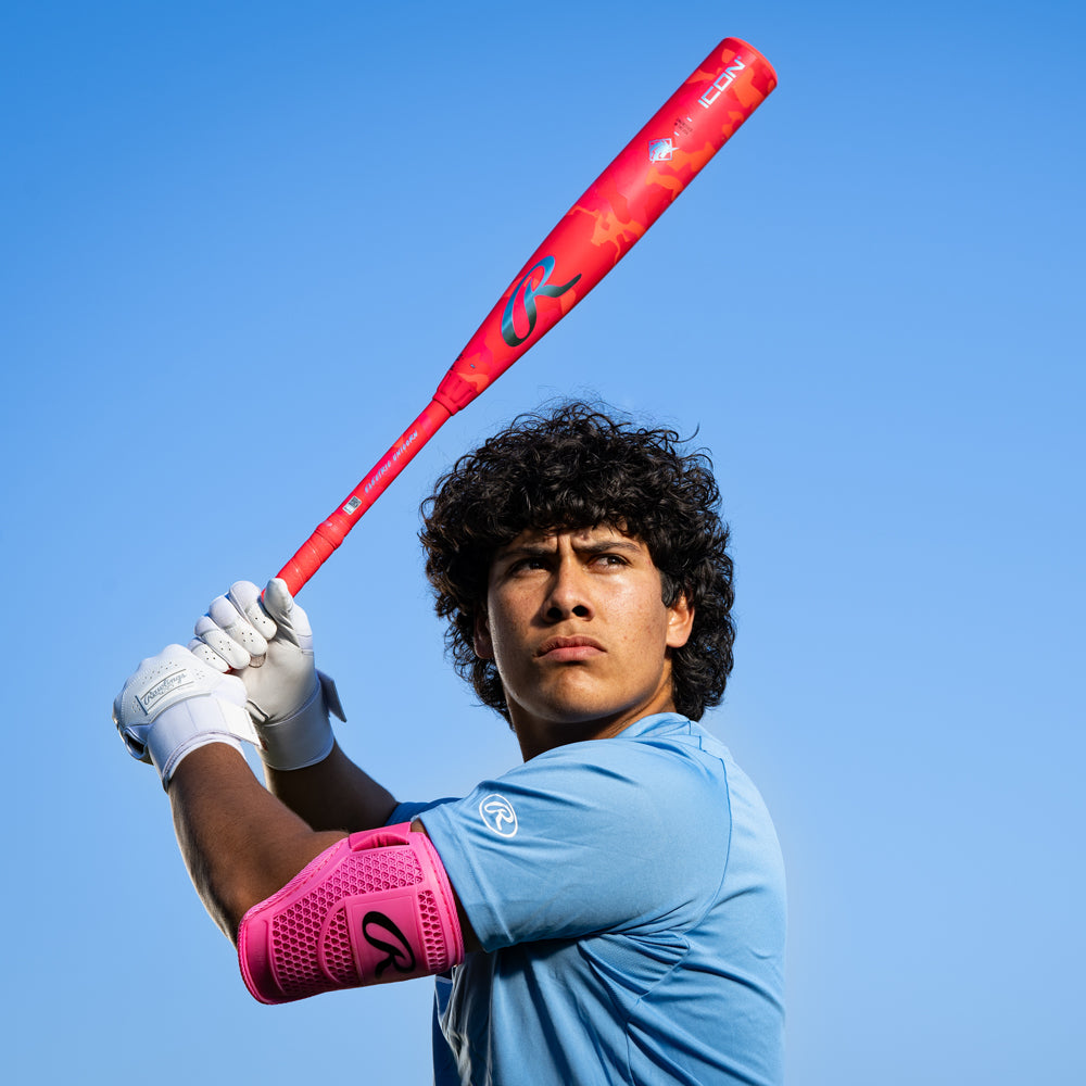 A young baseball player in a blue jersey holds the 2025 Rawlings Icon Electric Unicorn (-3) BBCOR Baseball Bat (RBB5IPK3), with white gloves and a pink arm guard, looking to the side under a clear blue sky.