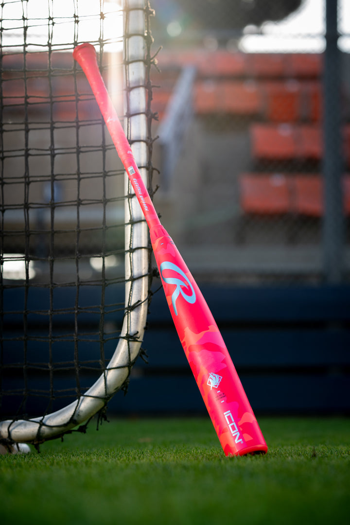 The 2025 Rawlings Icon Electric Unicorn (-3) BBCOR Baseball Bat rests against a net on a grassy field, its bright pink design with white and red accents standing out, with empty stadium seats in the background.
