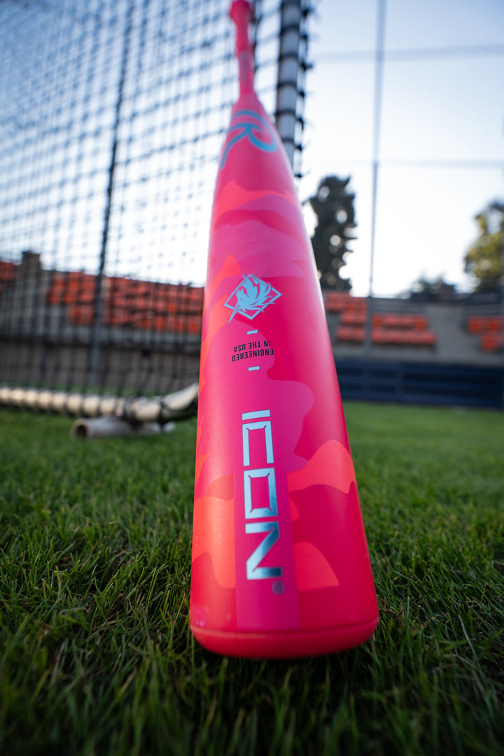 Close-up of a vibrant pink 2025 Rawlings Icon Electric Unicorn (-3) BBCOR Baseball Bat standing upright on green grass, with a net and empty orange stadium seats blurred in the background.