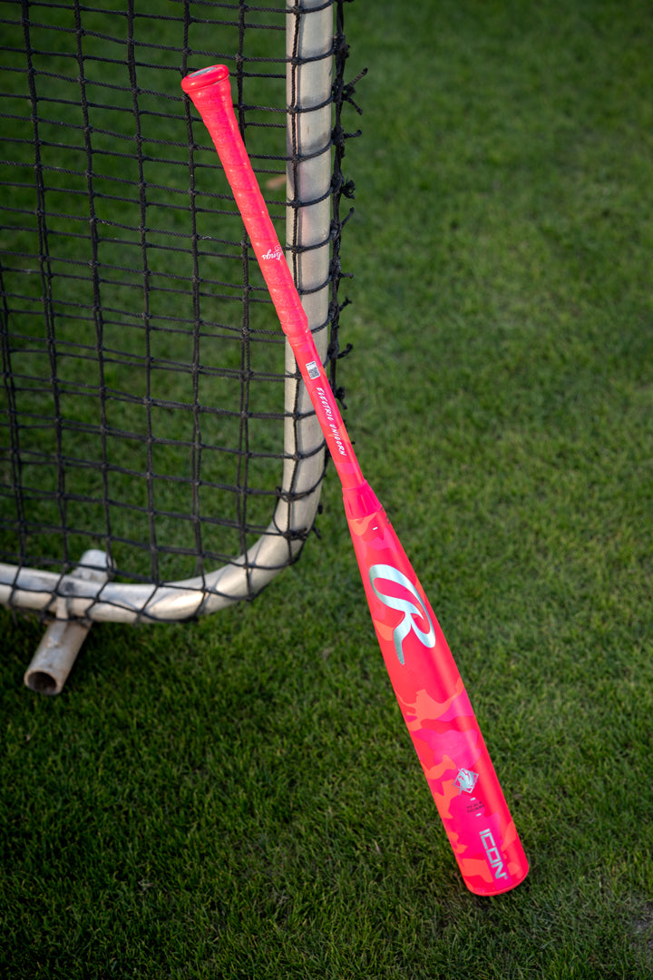 A 2025 Rawlings Icon Electric Unicorn (-3) BBCOR Baseball Bat (RBB5IPK3) with a pink camouflage design and large white “R” logo rests against a metal fence on green grass.