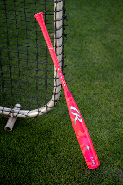 A 2025 Rawlings Icon Electric Unicorn (-3) BBCOR Baseball Bat (RBB5IPK3) with a pink camouflage design and large white “R” logo rests against a metal fence on green grass.