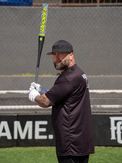 A bearded man in a black cap, black shirt, and white gloves stands on a sports field, holding a Dudley 2026 Lightning Legend Retro 2.0 Balanced SSUSA Bat (LLBSP2R) with a yellow and black design, looking over his shoulder at the camera.