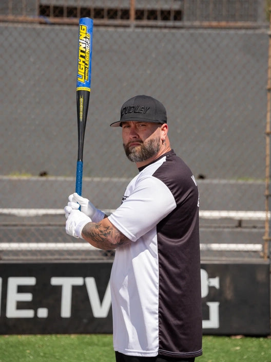 A man in a black and white jersey, black cap, and white gloves holds the Dudley 2026 Lightning Legend Retro 2.0 12" Endload SSUSA Senior Slowpitch Softball Bat (LLESP2R) upright on a sports field, with a chain-link fence and signage behind him.