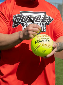 Wearing a bright red Dudley Softball Camps shirt, a person signs a yellow Dudley 18" Trophy Autograph Softball (4DAB18) by Dudley with a black marker; only their torso and hands are shown.