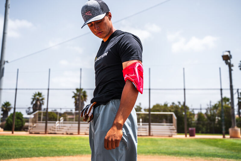 A baseball player on a field wears a black T-shirt, gray pants, cap, and the Easton Compact Batter Elbow Guard: ES37 by Easton, looking down with empty bleachers and palm trees in the background.