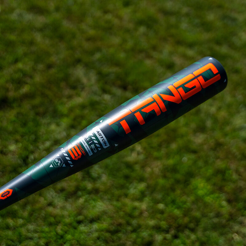A close-up of the 2025 Easton Tango (-8) 2 5/8" USA Baseball Bat: EUS5TNG8, with bold orange “TANGO” lettering on a dark green background, held above a grassy field—a great youth bat from Easton.