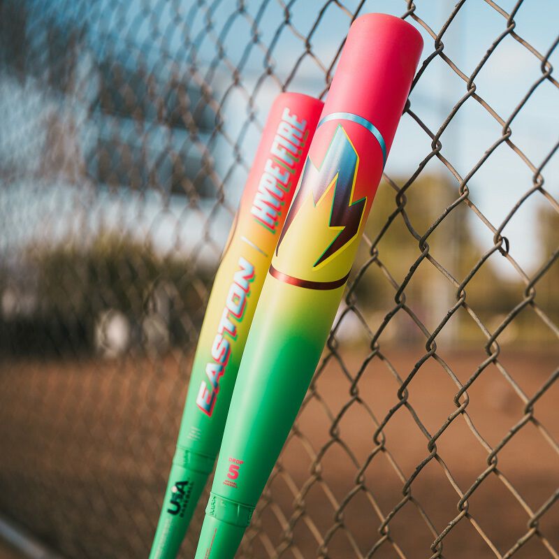 A group of baseball bats, including a 2026 Easton Hype Fire (-11) 2 5/8" USA Baseball Bat (EUS6HYP11), leaning on a fence.