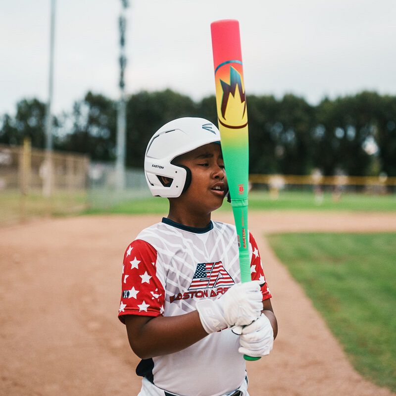 A young baseball player in a star-spangled jersey holds the 2026 Easton Hype Fire (-11) 2 5/8" USA Baseball Bat (EUS6HYP11) from Easton vertically on a field, wearing a helmet and gloves. Trees and a fence can be seen in the background.
