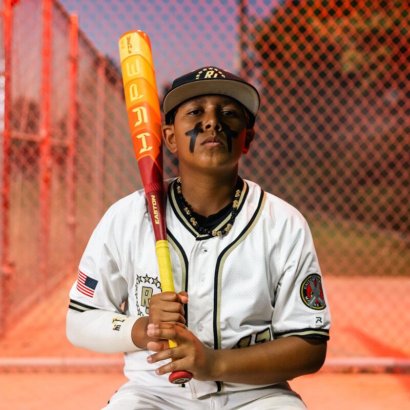 A young baseball player in a white uniform with black face paint sits by a chain-link fence on a sunny field, holding the 2025 Easton Hype Fire JBB (-10) 2 3/4" USSSA Baseball Bat: EJB5HYP10.