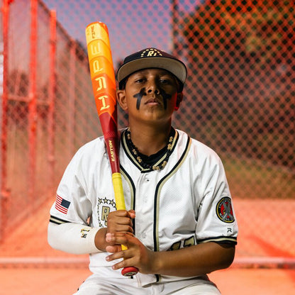 A young baseball player in a white uniform with black face paint sits by a chain-link fence on a sunny field, holding the 2025 Easton Hype Fire JBB (-10) 2 3/4" USSSA Baseball Bat: EJB5HYP10.