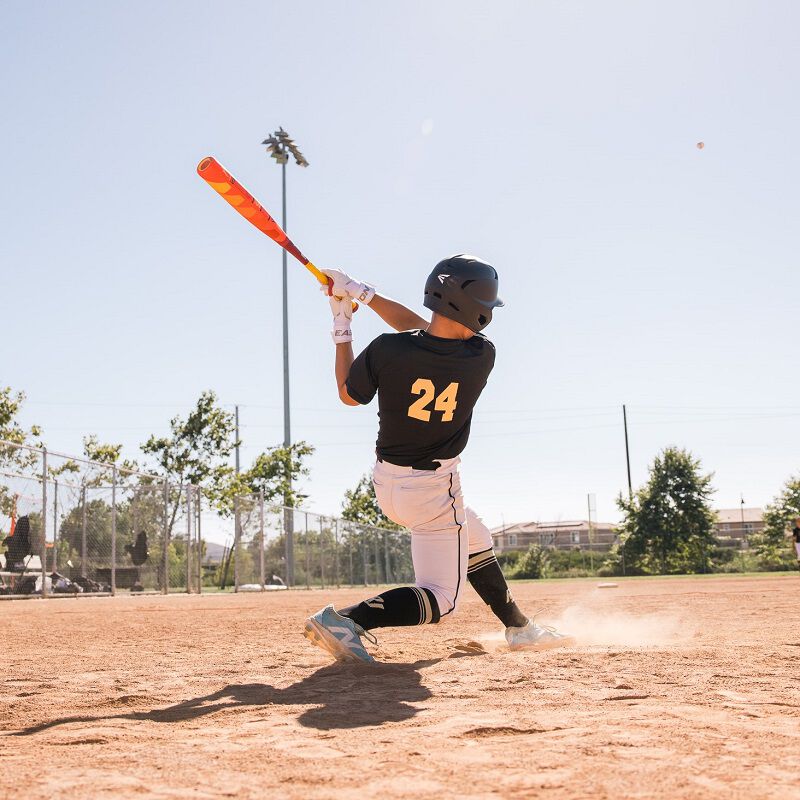 Wearing a black jersey with number 24, a baseball player swings the 2025 Easton Hype Fire JBB (-10) USSSA bat (EJB5HYP10) on a sunny field, launching the ball as dust rises, with trees and field lights in the background.