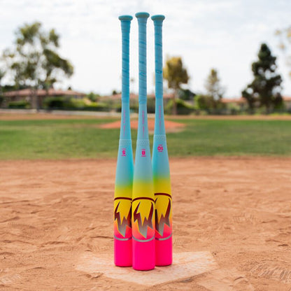 Three colorful bats, including the 2026 Easton Hype Fire (-5) 2 3/4" USSSA Baseball Bat by Easton, stand upright on the field. Their blue handles and pink/yellow gradient barrels contrast with the grass, trees, and clear sky.