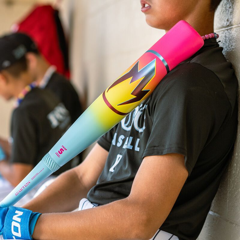 A baseball player in black jersey and white pants leans against a dugout wall, holding the 2026 Easton Hype Fire (-8) 2 3/4" USSSA Baseball Bat (EUT6HYP8) by Easton over his shoulder. Another player is visible in the blurred background.