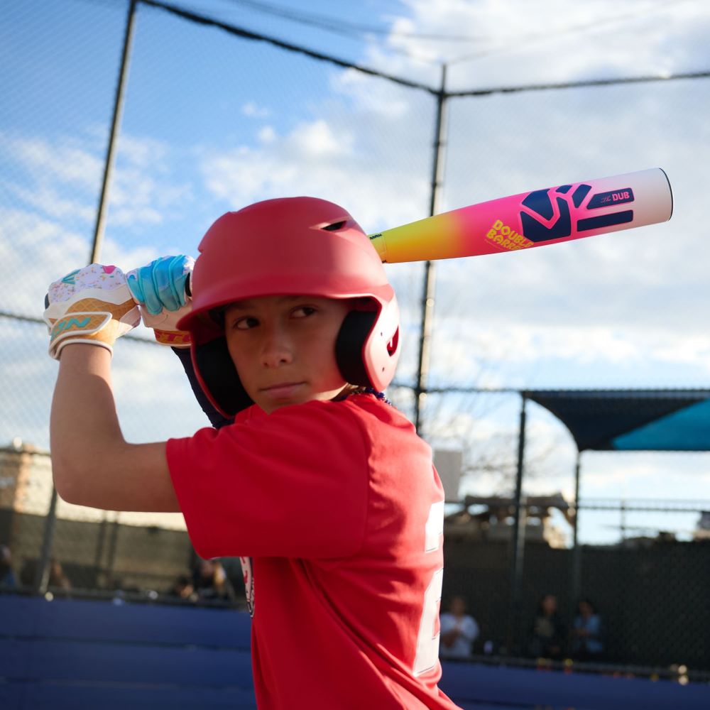 A young baseball player in a red shirt and helmet grips the 2026 Easton Dub (-10) 2 3/4" USSSA Baseball Bat: EUT6DUB10, ready to swing on an outdoor field with a blue sky and chain-link fence in the background.