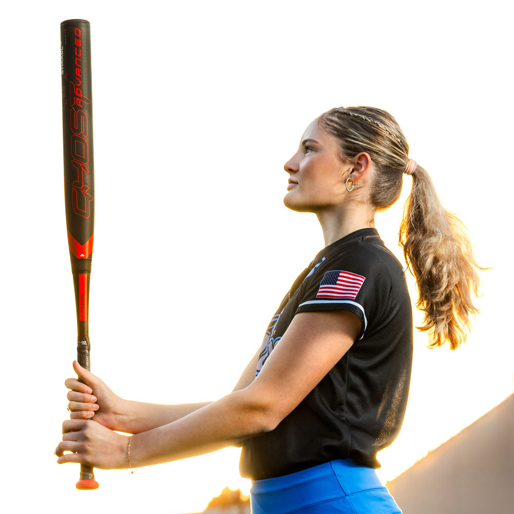 A young woman with a ponytail holds the 2024 Easton Ghost Advanced (-9) Fastpitch Softball Bat (EFP4GHAD9), wearing a black jersey with an American flag patch on the sleeve, standing outdoors against a bright sky.