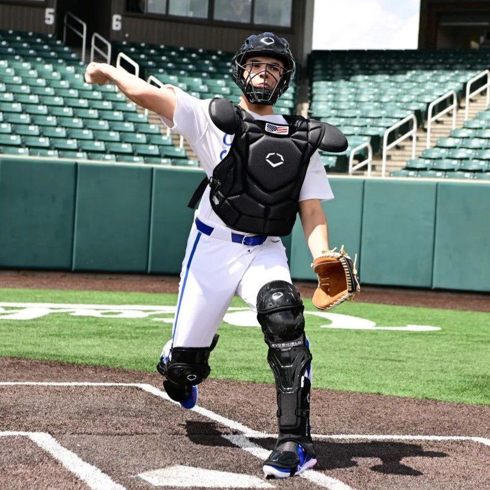 A baseball catcher in full gear throws a ball on a field, with green stadium seats and railings visible in the background.