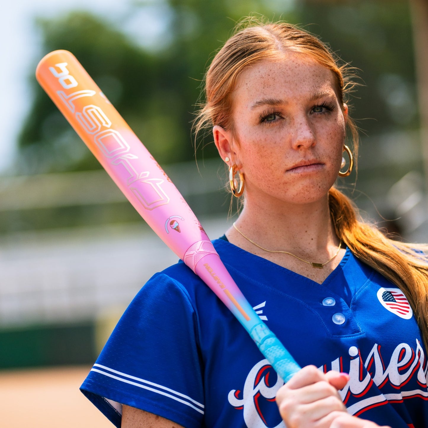 A young woman in a blue softball jersey confidently poses outdoors with the 2025 Easton Ghost OG Rainbow Sherbet (-11) Fastpitch Softball Bat (EFP5GHRS11) by Easton resting on her shoulder, trees and field softly blurred behind her.
