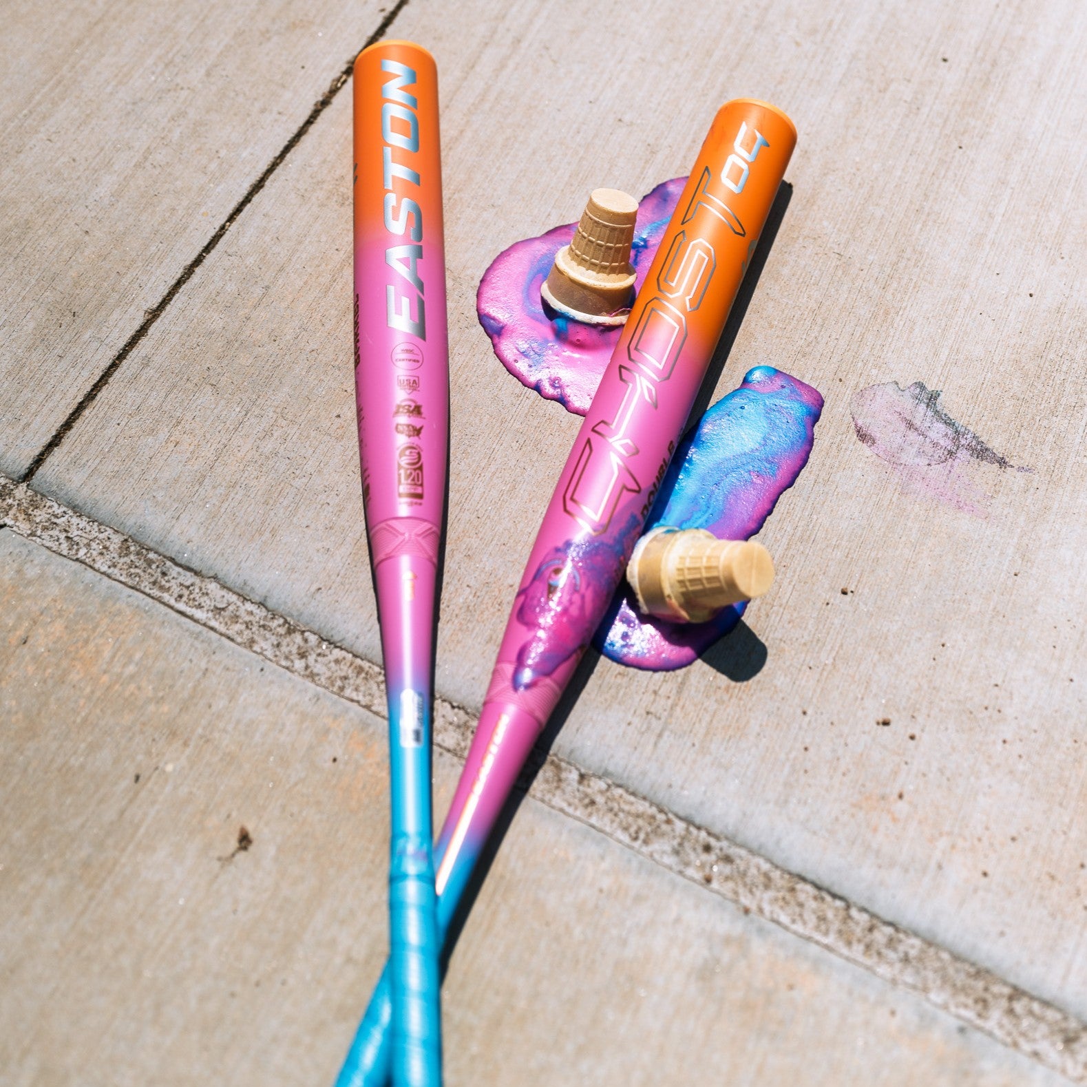 Two vibrant Easton Ghost OG Rainbow Sherbet (-11) Fastpitch Softball Bats rest on a sidewalk beside two melted ice cream cones with pink and purple puddles, highlighting the colorful 2025 design and advanced Double Barrel construction.
