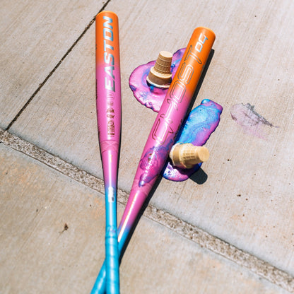 Two vibrant Easton Ghost OG Rainbow Sherbet (-11) Fastpitch Softball Bats rest on a sidewalk beside two melted ice cream cones with pink and purple puddles, highlighting the colorful 2025 design and advanced Double Barrel construction.