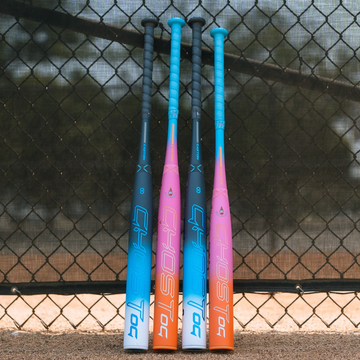 Three vibrant 2025 Easton Ghost OG Rainbow Sherbet (-11) Fastpitch Softball Bats stand upright by a baseball field fence, displaying “GHOST,” Double Barrel construction, and Sonic Comp technology. Dirt and grass are seen behind them.