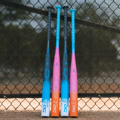 Three vibrant 2025 Easton Ghost OG Rainbow Sherbet (-11) Fastpitch Softball Bats stand upright by a baseball field fence, displaying “GHOST,” Double Barrel construction, and Sonic Comp technology. Dirt and grass are seen behind them.