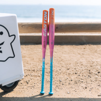 Two 2025 Easton Ghost OG Rainbow Sherbet (-11) Fastpitch Softball Bats with pink, orange, and blue accents stand upright on sandy ground near a wooden fence by the ocean. A white Ghost graphic box is partially visible. Features Double Barrel construction.