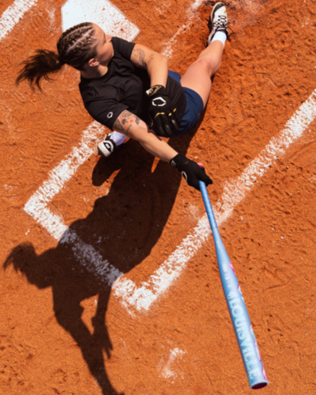 With her hair in braids, a softball player swings a blue Louisville Slugger Kryo One (-10) Fastpitch Softball Bat (WBL4273010) by Louisville Slugger on a dirt field, casting a shadow across home plate.