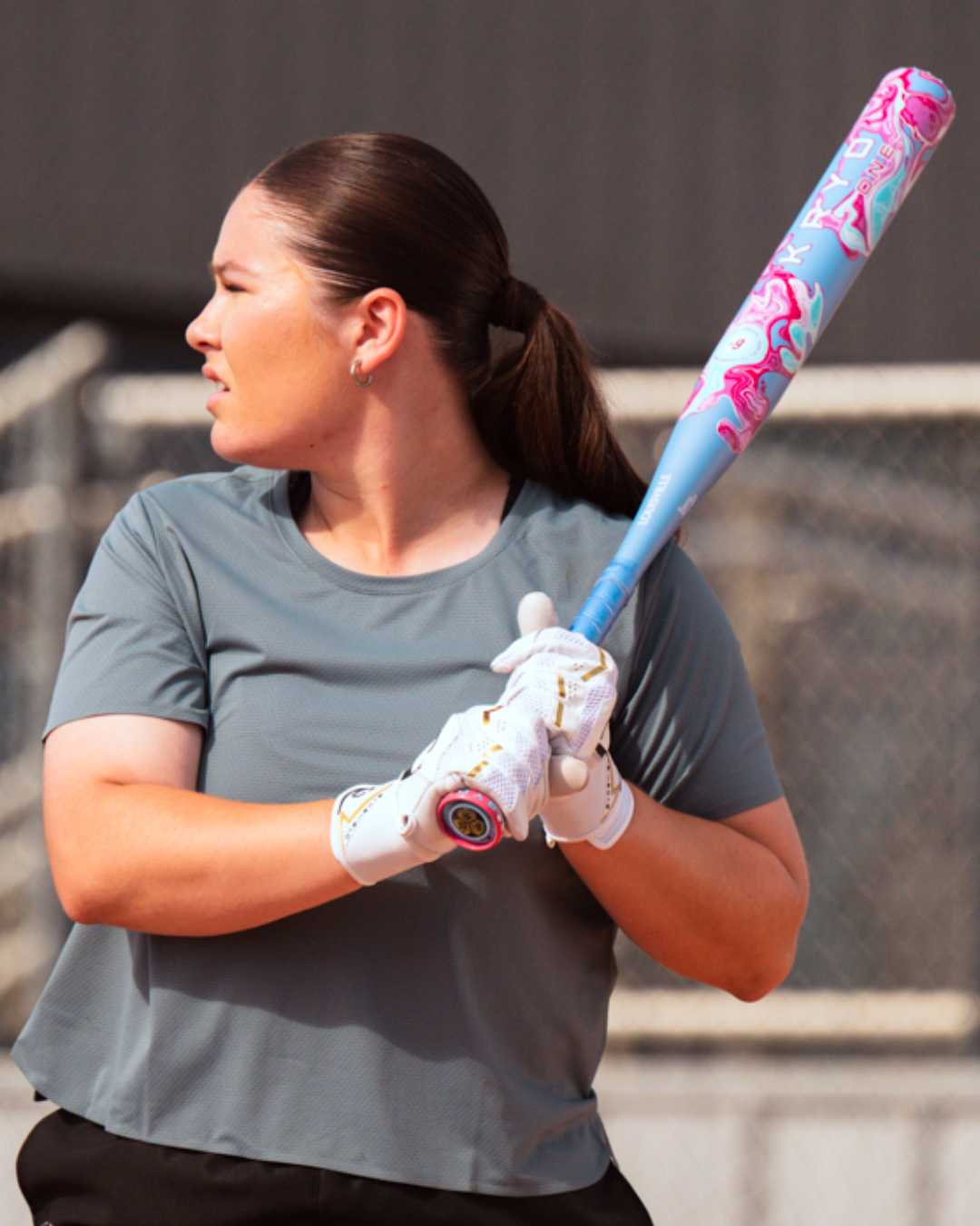 A woman with a ponytail, dressed in a gray shirt, stands in front of a chain-link fence holding the 2027 Louisville Slugger Kryo One (-8) Fastpitch Softball Bat (WBL4275010), poised to swing as she looks to her left.