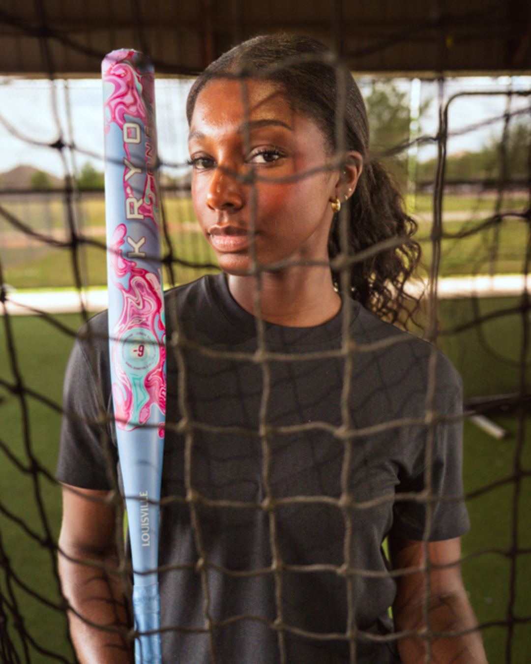A woman in a black shirt holds a vibrant 2027 Louisville Slugger Kryo One (-11) Fastpitch Softball Bat (WBL4272010), standing behind a net on a green indoor field and looking directly at the camera.