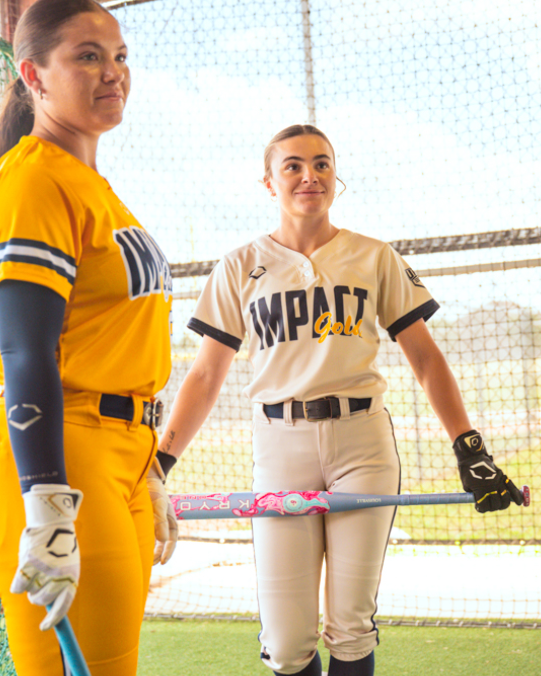 Two softball players in Impact Gold uniforms stand indoors. One wears yellow, the other white; both hold gloves and swing their bats—a fastpitch softball bat and the Louisville Slugger Kryo One (-10) Fastpitch Softball Bat (WBL4273010)—against a backdrop of netted walls and streaming sunlight.