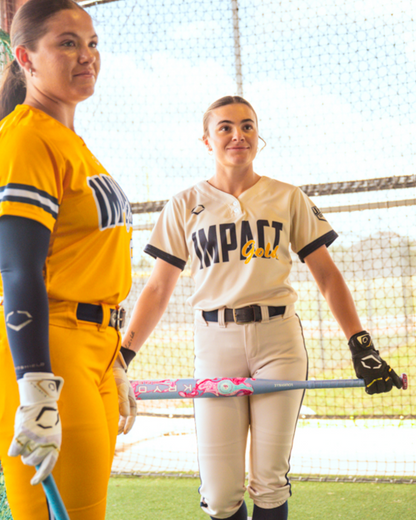 Two softball players in Impact Gold uniforms stand indoors. One wears yellow, the other white; both hold gloves and swing their bats—a fastpitch softball bat and the Louisville Slugger Kryo One (-10) Fastpitch Softball Bat (WBL4273010)—against a backdrop of netted walls and streaming sunlight.