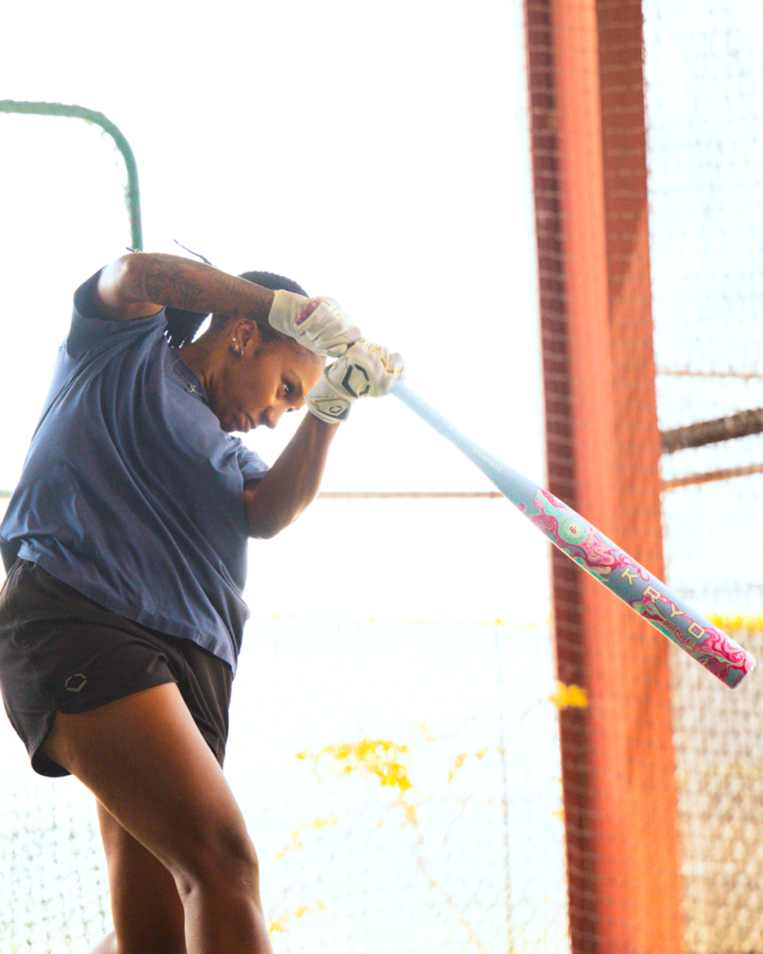 A person wearing a blue shirt and shorts prepares to swing the Louisville Slugger 2027 Kryo One (-11) Fastpitch Softball Bat (WBL4272010), a colorful one-piece composite bat, inside a batting cage.