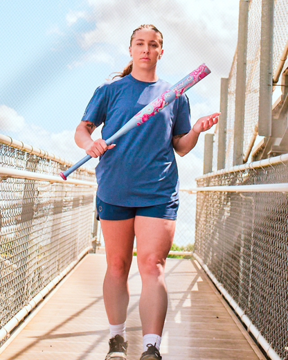 A woman in a blue shirt and shorts stands confidently on a bridge holding the 2027 Louisville Slugger Kryo One (-10) Fastpitch Softball Bat (WBL4273010), with a blue sky and clouds in the background.