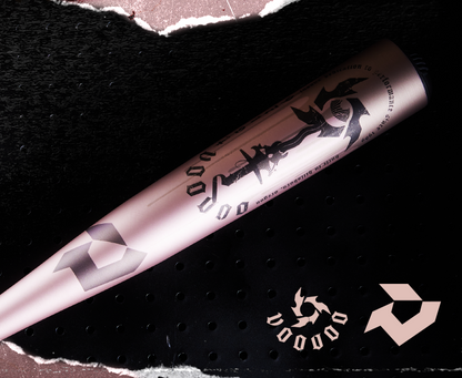 The 2025 DeMarini Voodoo One Rose Gold (-3) BBCOR Baseball Bat (WBD2628010) features rose gold color, black graphics, and VOODOO branding, shown on a perforated black surface beside a matching logo and VOODOO text design.