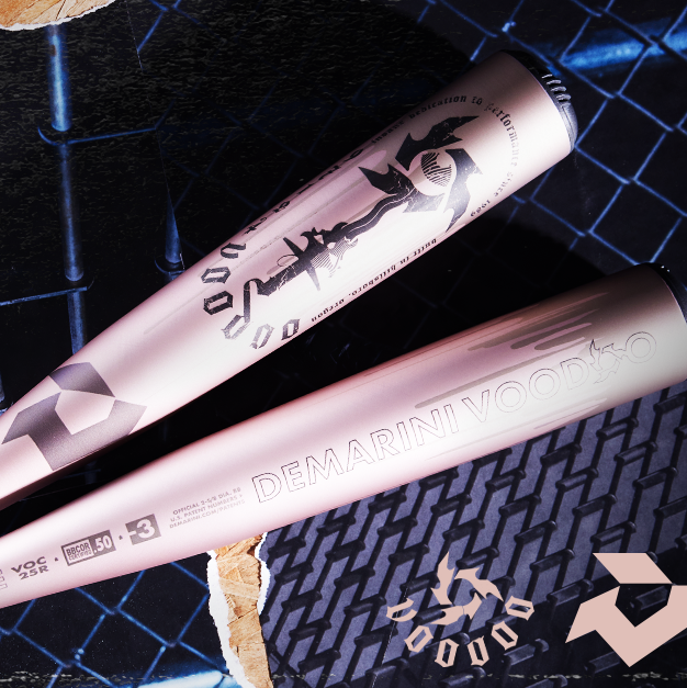 Two metallic DeMarini Voodoo baseball bats with bold branding lie on a dark textured metal surface, illuminated by dramatic lighting. The DeMarini logo and Voodoo text are clearly visible on the bats.