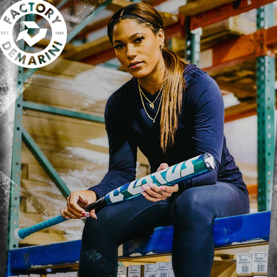 A woman in athletic wear sits on a warehouse shelf, holding a teal 2026 DeMarini Lotus (-11) Fastpitch Softball Bat: WBD2605010 (DEMO). She looks at the camera seriously. The DeMarini logo appears in the top left corner.