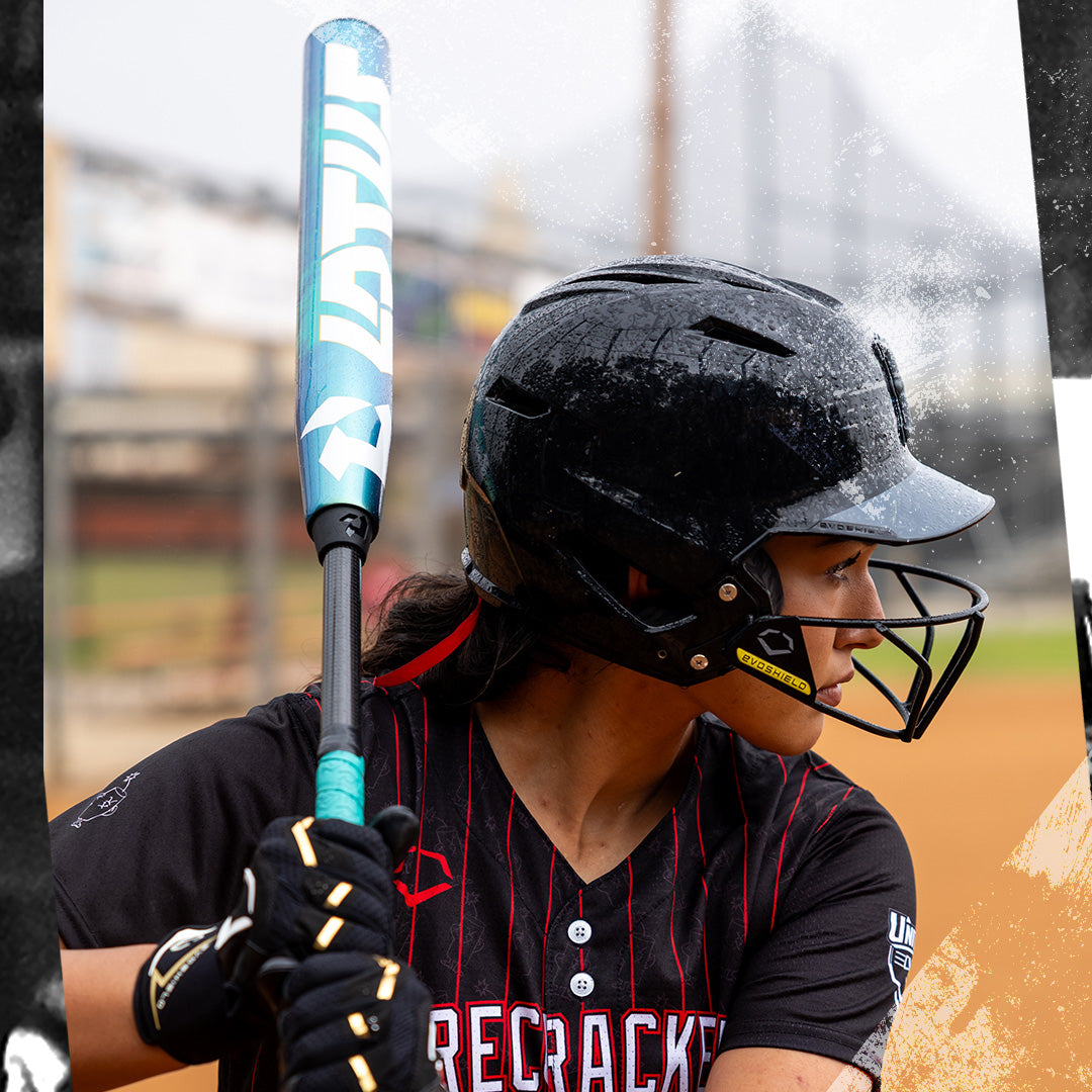 A softball player in a black helmet and jersey with red stripes grips the 2026 DeMarini Lotus (-11) Fastpitch Softball Bat: WBD2605010 (DEMO), preparing to bat on a dirt field with a blurred fence and building in the background.