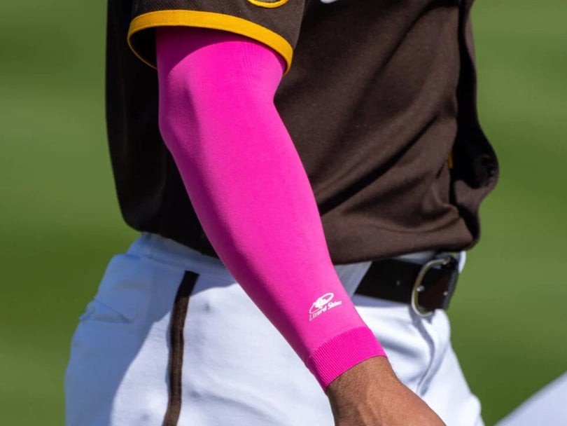 A baseball player wearing a brown jersey and white pants is shown from the waist up, with a bright pink compression sleeve on their right arm. The background is blurred green grass.