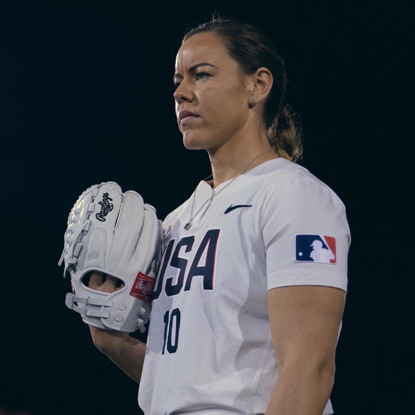 A focused female softball player in a white USA jersey with the number 10 and an MLB patch holds a white glove, standing against a dark background.