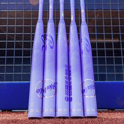 Five purple softball bats with Rawlings branding stand upright in a row against a metal fence on brown dirt, with a blue padded barrier behind—ideal for players seeking their next bat like the 2027 Rawlings Halo (-9) Fastpitch Softball Bat: RFP7HALO9.
