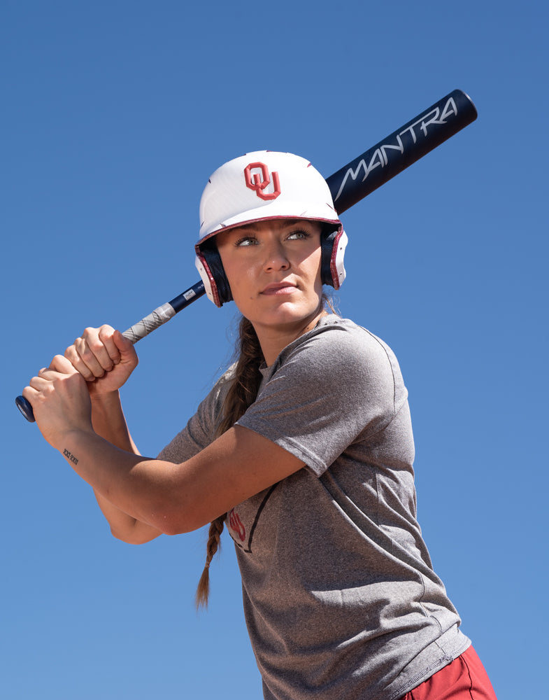 A softball player in a gray shirt and white helmet with an OU logo holds the 2025 Rawlings Mantra (-10) Fastpitch Softball Bat: RFP4M10 (DEMO), ready to swing under a clear blue sky.