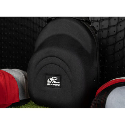 The Lizard Skins Hat Guardian: HATGD100, a black hard-shell case, stands upright on artificial grass with caps around it and a black pegboard backdrop. This crush-proof case from Lizard Skins protects your hat collection.
