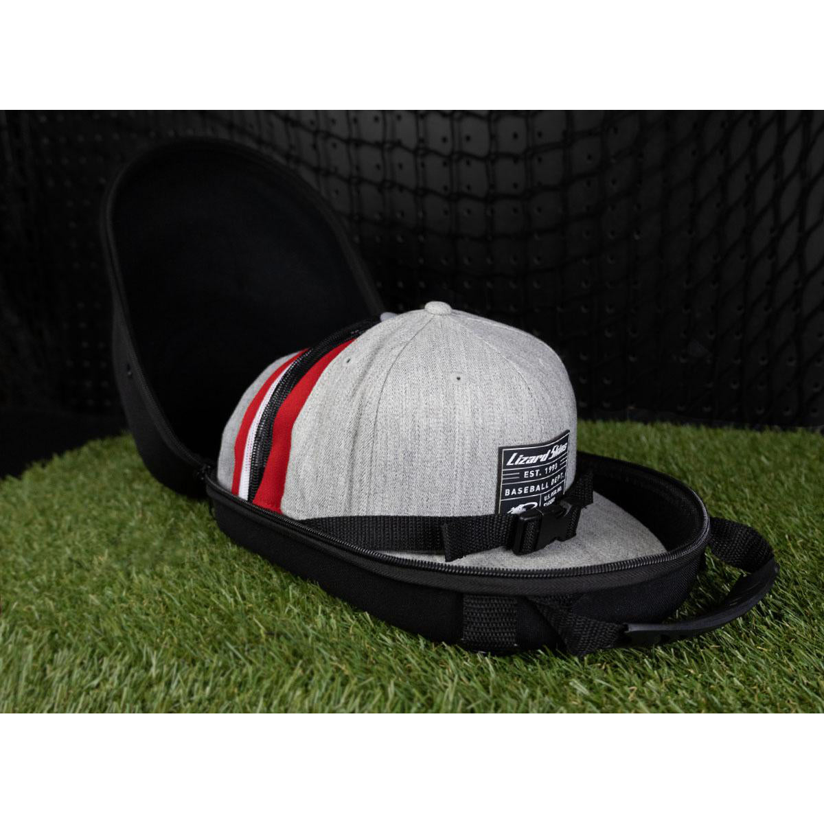 A gray baseball cap with red and black stripes sits inside a Lizard Skins Hat Guardian: HATGD100 crush proof case by Lizard Skins, open on artificial grass against a black textured background.