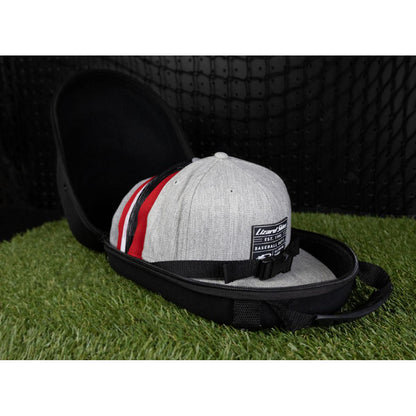 A gray baseball cap with red and black stripes sits inside a Lizard Skins Hat Guardian: HATGD100 crush proof case by Lizard Skins, open on artificial grass against a black textured background.