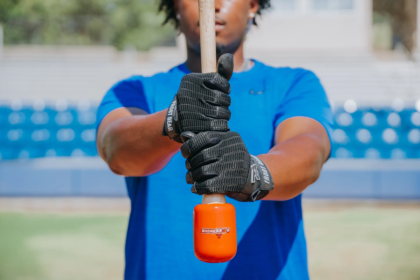 A person in black gloves and a blue shirt grips a bat with the orange Hitting Knob Bat Weight by Hitting Knob, standing on a field with empty stadium seats to help improve bat speed.
