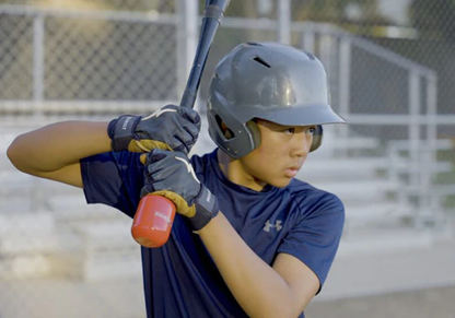 A young baseball player wearing a helmet and gloves grips a bat featuring the Hitting Knob Bat Weight by Hitting Knob, ready to swing near empty bleachers and a chain-link fence on the baseball field.