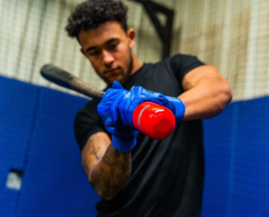 A baseball player in blue gloves grips a bat outfitted with the red Hitting Knob Bat Weight by Hitting Knob, preparing to swing in an indoor training facility.